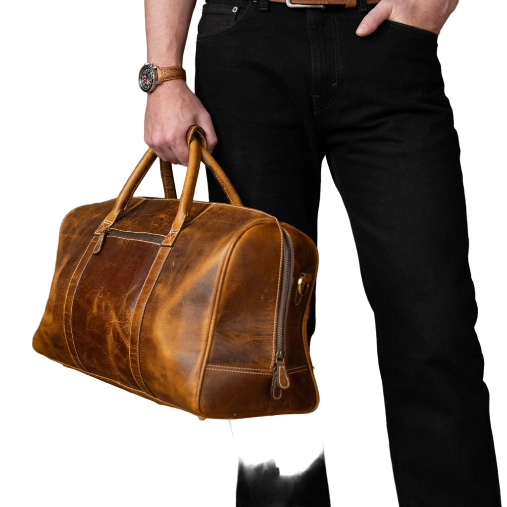 "Person carrying The Wayfarer Bag by the top handle — cognac full-grain leather duffle bag by NAB Leather, ideal for weekend travel"