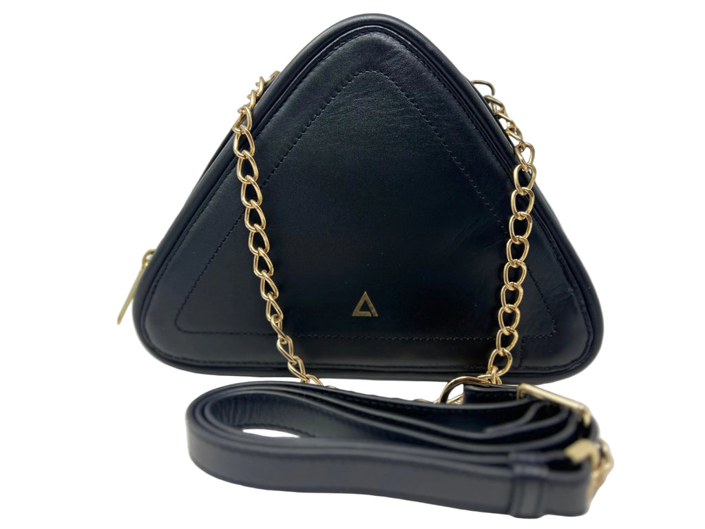 Front view of the Annick Levesque Kiana triangular black leather evening bag with a gold chain strap, a small triangle logo on the front, and a coiled black leather shoulder strap lying in front against a white background.