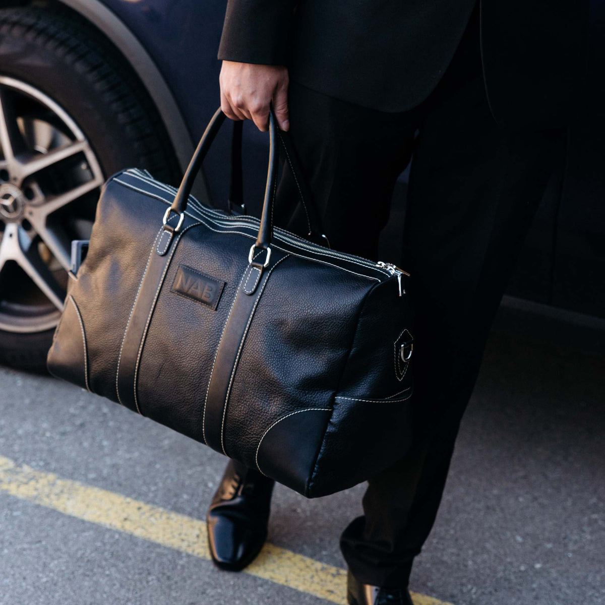 "Person carrying The Shield Duffle in black at their side outdoors — full-grain leather carry-on duffle bag made in Canada by NAB Leather"