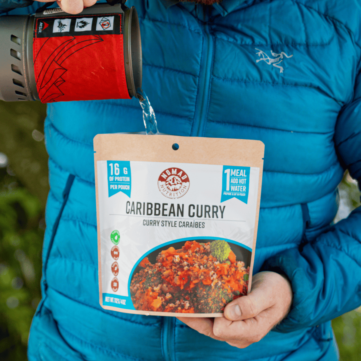 A person in a blue puffer jacket holds a package of Namu Caribbean Curry and pours hot water from a red camping mug into the pouch. The package features a kraft paper design with a photo of the prepared curry dish.