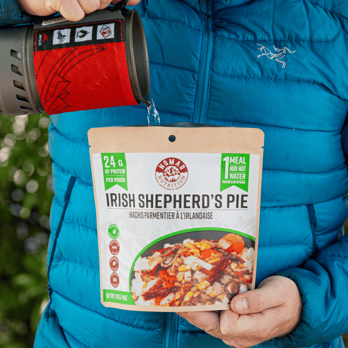 A person in a bright blue puffer jacket holds a pouch of Fingal's Mountain Shepherd's Pie while pouring hot water from a red and gray camping mug into the package.