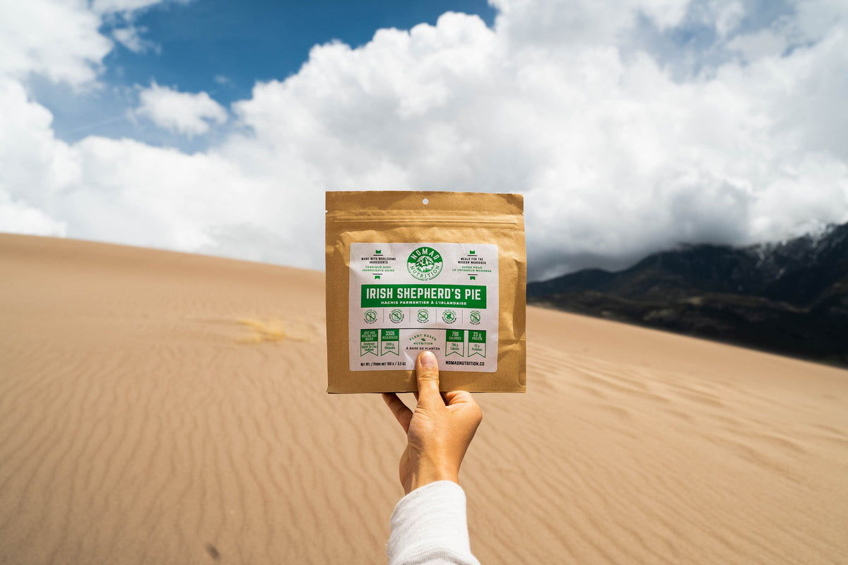 Hand holding a package labeled 'Irish Shepherd's Pie' against a desert landscape with mountains and blue sky.
