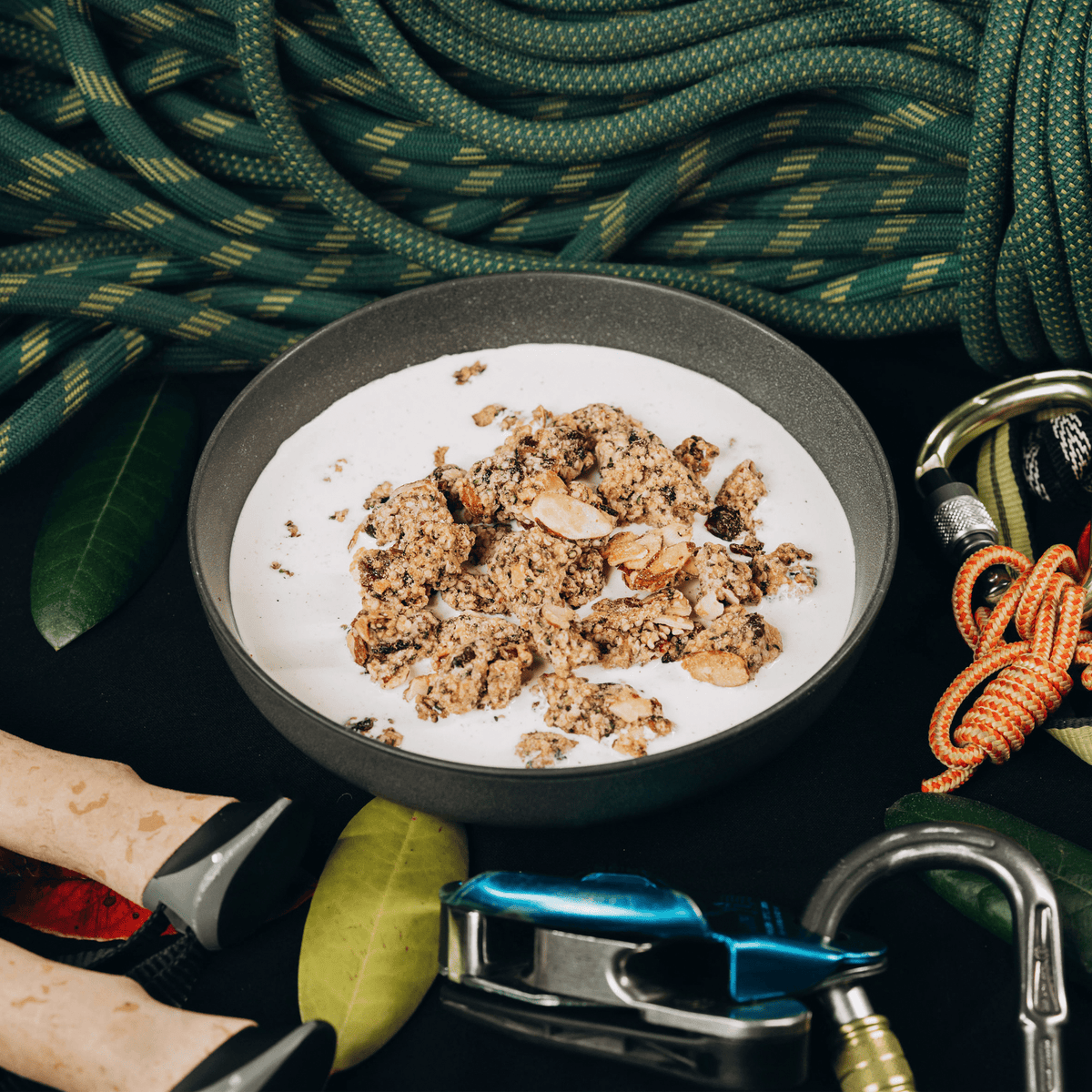 Bowl of granola with milk surrounded by climbing equipment on a dark surface